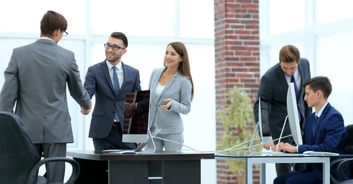 A group of people in an office dressed in business attire. Two men wearing glasses are shaking hands.