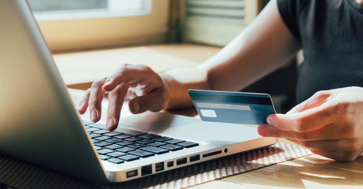 A person holding a credit card and typing on their laptop. There is a window next to them, and the sun is shining.