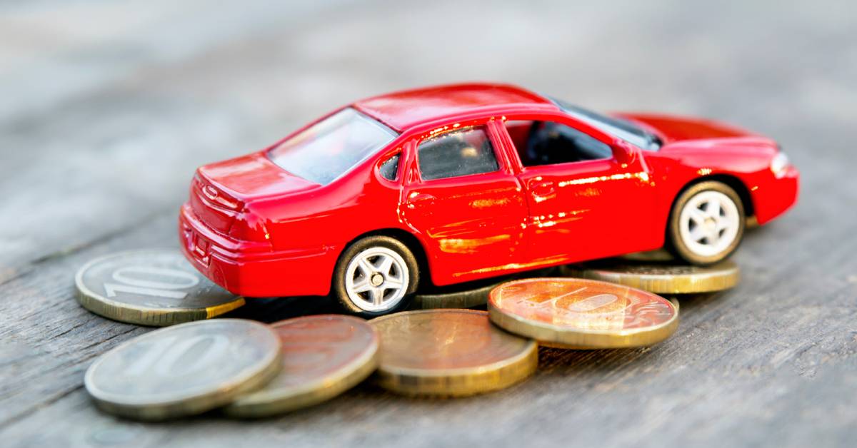 A miniature bright red sedan lying on top of an unstacked pile of coins. The car and the coins are on a wooden surface.